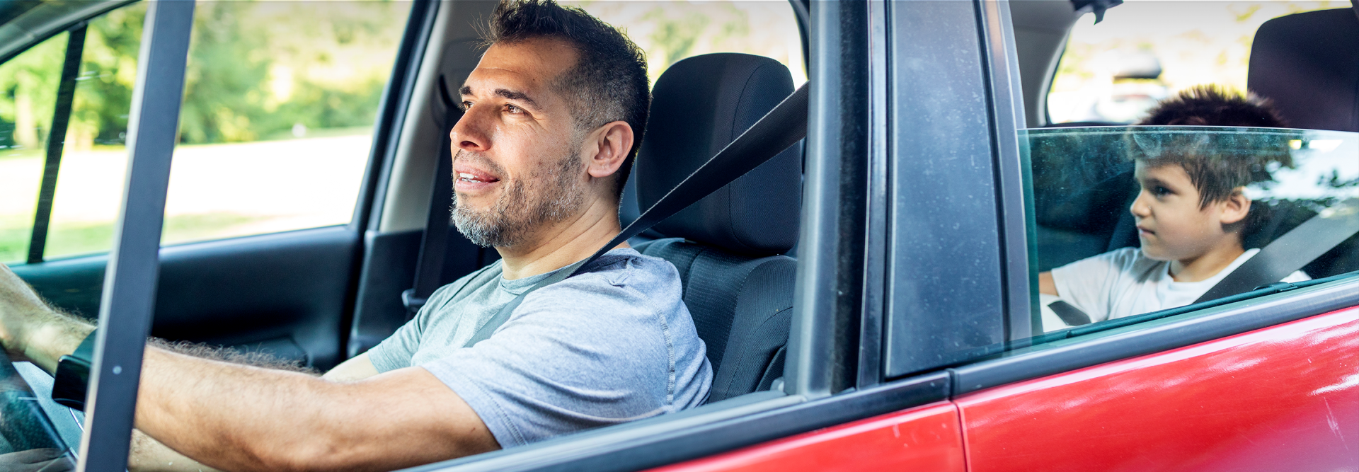 Photo of father driving with his seat belt on looking in his rearview mirror at his young son who has his seat belt on in the backseat and is holding a soccer ball